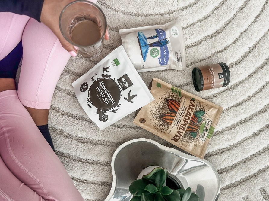 Lattialla risti-istunnassa olevat jalat ja kdess lasillinen ashwagandhajuomaa, lattialla mys Healthwell Ashwagandha-jauhe Luomu, Celtic Havsalt ja Rawpowder kaakaojauhe -pussit ja Urtekrm kanelipurkki sek viherkasvi.